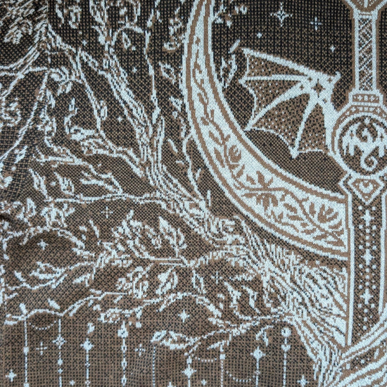 Close-up of a brown woven textile with white embroidered or printed fantasy-style artwork, including a crescent moon, bat, stars, and abstract line motifs.