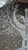 Close-up of a brown woven textile with white embroidered or printed fantasy-style artwork, including a crescent moon, bat, stars, and abstract line motifs.